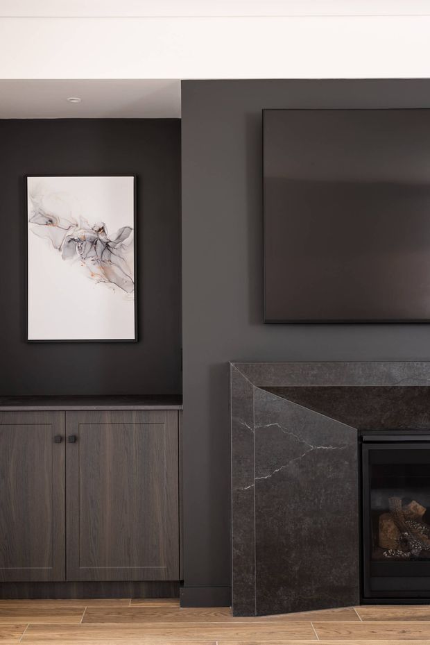 A moody contemporary living room feature wall in charcoal tones, with a wall-mounted television, abstract artwork, dark timber cabinetry, and a dark stone-clad fireplace surround. Warm timber flooring contrasts with the dark built-in joinery and fireplace.
