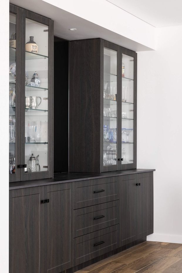 A built-in dark timber display and storage cabinet spans the wall, with glass-fronted upper cabinets showcasing glassware and serving pieces. The lower section has solid-panel doors and drawers with black hardware, set against white walls and a warm timber floor. The clean, restrained joinery gives the space a refined contemporary appearance.