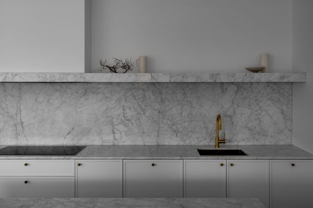 A refined minimalist kitchen with pale grey cabinetry, a dark stone benchtop, a black inset sink and a gold-toned tap. A dramatic veined marble splashback rises to a full-width floating shelf, with sparse decorative objects above. The subdued palette and low lighting create a calm, elegant atmosphere.