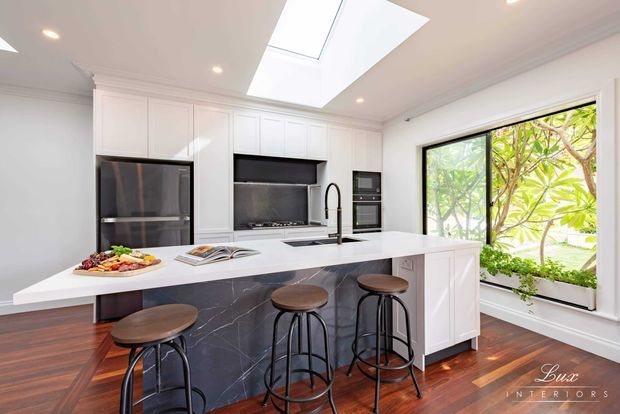 Bright contemporary kitchen with white shaker-style cabinetry, a large waterfall island clad in dark veined stone, integrated stainless-steel appliances, skylight, recessed lighting, and a large garden-facing window. The island includes seating for four, and the room has polished dark timber flooring with restrained black accents.