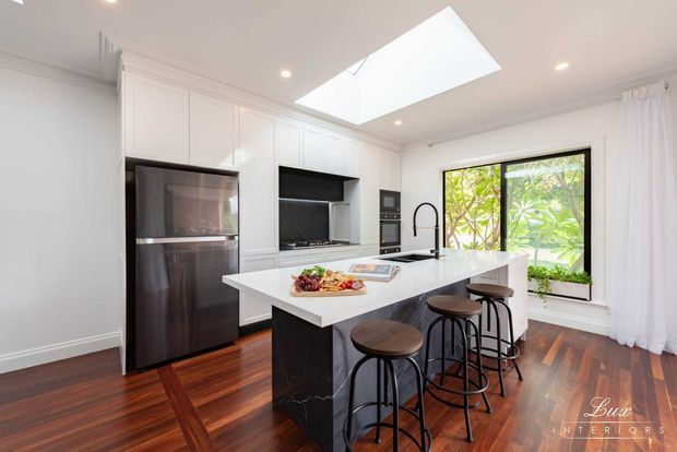 Bright contemporary kitchen with a large white island and dark waterfall-edge base, four bar stools, integrated white cabinetry, black appliances, timber flooring, a skylight, and a large garden-facing window with sheer curtains.