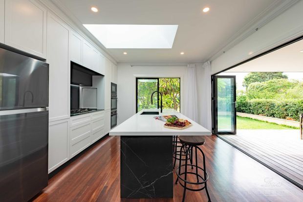 Bright contemporary kitchen with white floor-to-ceiling cabinetry, integrated appliances, a large white island with a black marble-look base, and black bar stools. Dark timber flooring contrasts with the light cabinetry, while a large skylight and expansive glazed folding or stacking doors provide abundant natural light and connect the kitchen to a timber outdoor deck and landscaped garden.