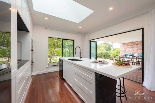 Bright contemporary white kitchen with a large island, integrated cabinetry, dark timber flooring, skylight, and expansive opening to an outdoor entertaining area. The island includes a sink and seating, while garden views and a brick exterior wall are visible beyond the glazing.