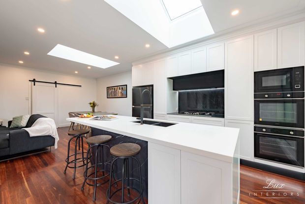 Bright open-plan kitchen with a large white island and seating for four, white shaker-style cabinetry, integrated black appliances, dark stone-look splashback, skylights, and warm timber flooring. A living area with a sofa is visible to the left, separated by a sliding barn-style door.