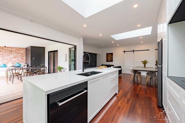 Bright open-plan kitchen and dining area with a large white island, integrated sink and dark dishwasher, white cabinetry, polished dark timber flooring, skylights, and black-framed glazed doors opening to a dining terrace. A brick feature wall and adjacent dining area are visible beyond the kitchen.