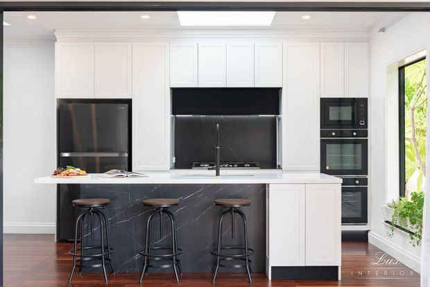 Bright, refined contemporary kitchen with white shaker-style cabinetry, a large island clad in dark veined stone, integrated appliances, and three black metal stools. The room features timber flooring, recessed lighting, a skylight, and generous windows overlooking greenery.
