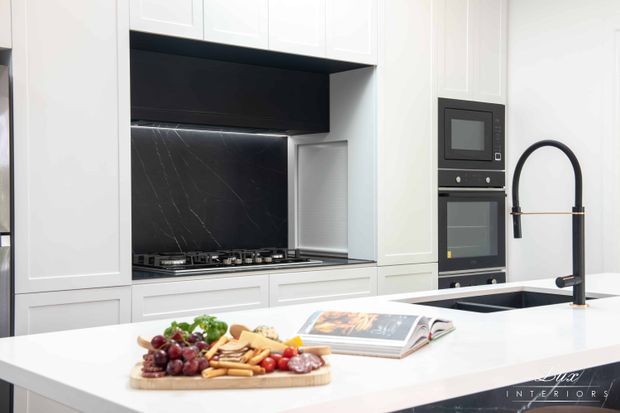 Bright, refined contemporary kitchen featuring white panelled cabinetry, a large white island benchtop, integrated black appliances, a black marble-look splashback, gas cooktop, and a distinctive matte-black gooseneck tap. An open cookbook and grazing board sit on the island.