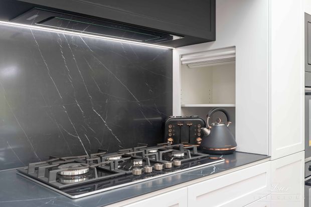 Close-up of a contemporary kitchen cooking zone with a six-burner gas cooktop, dark cabinetry, a dramatic black marble-look splashback, and a matching dark rangehood. White lower cabinetry and a built-in open appliance cupboard provide contrast, while a dark kettle sits on the cooktop.