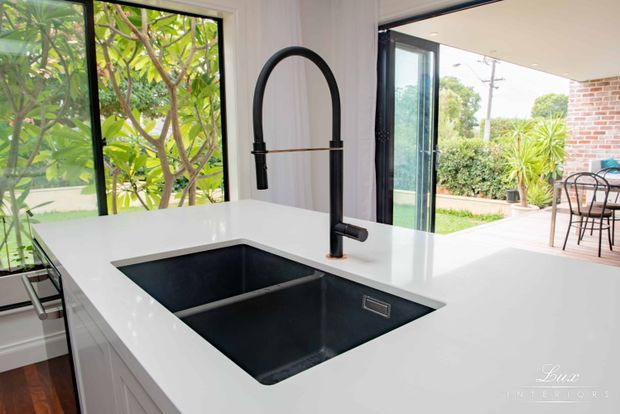 Bright contemporary kitchen focused on a large white island with an integrated black double-bowl sink and tall matte-black gooseneck tap. Full-height black-framed glazing opens views to a landscaped patio, with white cabinetry, light walls, timber flooring, and a small outdoor dining area visible beyond.
