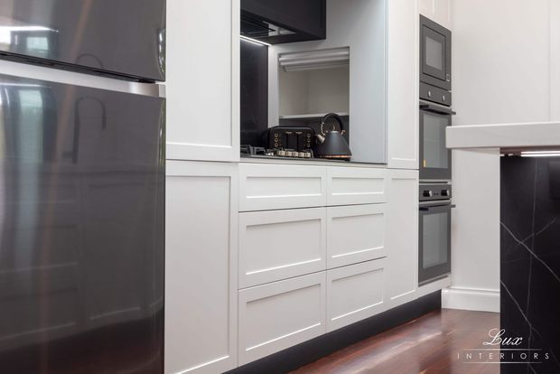 A compact contemporary kitchen featuring white recessed-panel cabinetry, dark integrated appliances, a stainless-steel refrigerator, and a black marble-look island side with white veining. Warm timber flooring contrasts with the monochromatic cabinetry and appliances.