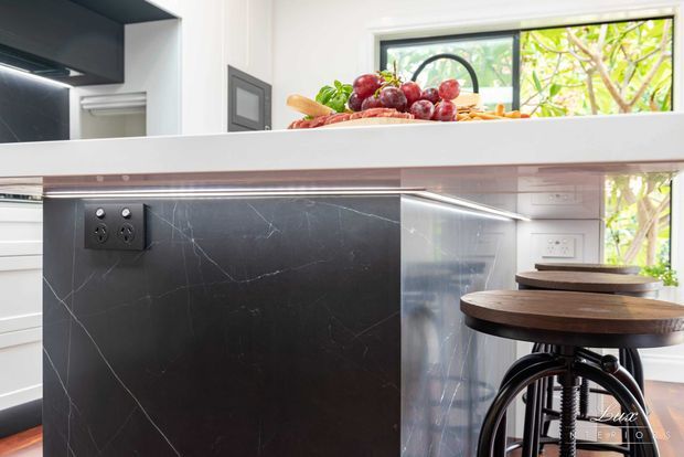 Close-up view of a contemporary kitchen island and breakfast bar. The island features a dark marble-look slab panel with integrated power outlets, a white benchtop, under-bench lighting, and timber-topped industrial-style stools. White cabinetry and a large window provide a bright backdrop, with produce arranged on the counter.
