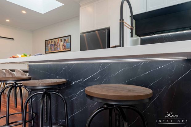 Contemporary kitchen island with a dark marble-look waterfall or panelled front, white cabinetry, integrated appliances, a skylight, and a raised light benchtop. Three round timber-topped black metal bar stools create a casual seating area. A tall arched black tap and under-cabinet lighting add strong modern detailing.