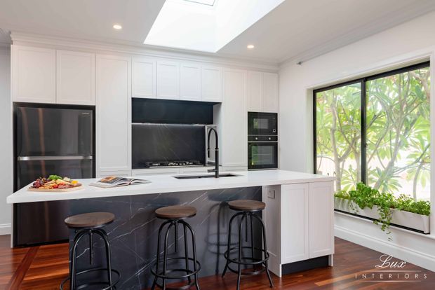 Bright, refined kitchen with white Shaker-style cabinetry, a large island with a dramatic dark veined stone waterfall end, integrated black appliances, skylight, and black-framed glazing overlooking greenery. Three stools create casual island seating, while warm timber flooring adds contrast.