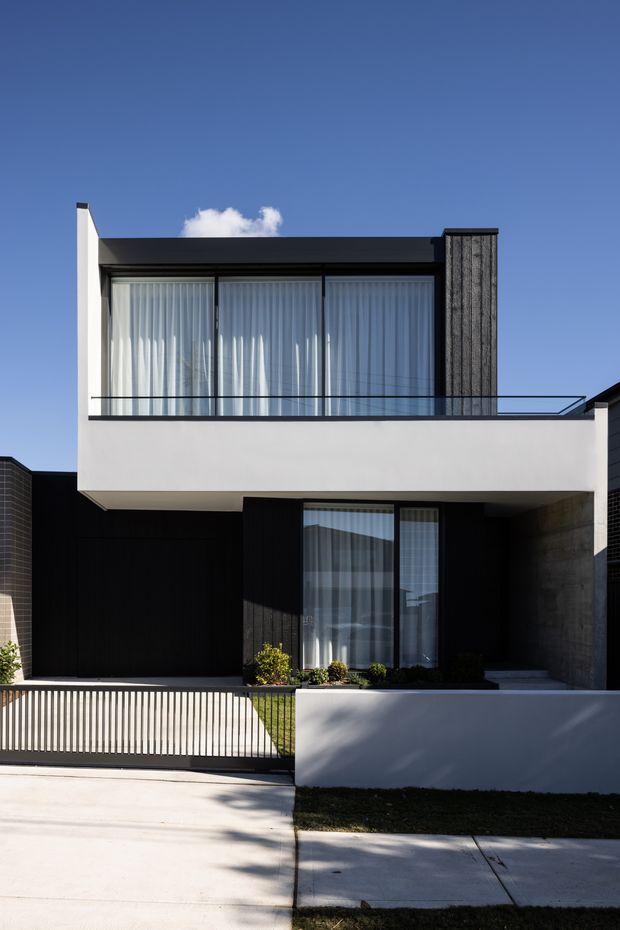 Street-facing view of a contemporary two-storey residence with a crisp rectilinear form, flat roof, white rendered upper volume, dark cladding, full-height glazed doors and windows, and a glass-balustraded upper balcony. A vertical textured dark feature wall, recessed ground-floor entry, landscaped frontage, and simple metal driveway gate complete the facade.