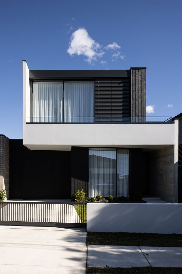 A front exterior view of a contemporary two-storey residence with a rectilinear flat-roof form. The facade combines smooth white rendered walls, dark vertical cladding, extensive upper-level glazing with sheer curtains, black horizontal louvres, and a glass balustrade. A recessed entry and garage area sit beneath the cantilevered upper floor, with a minimalist front fence, driveway, lawn, and small landscaped planting.