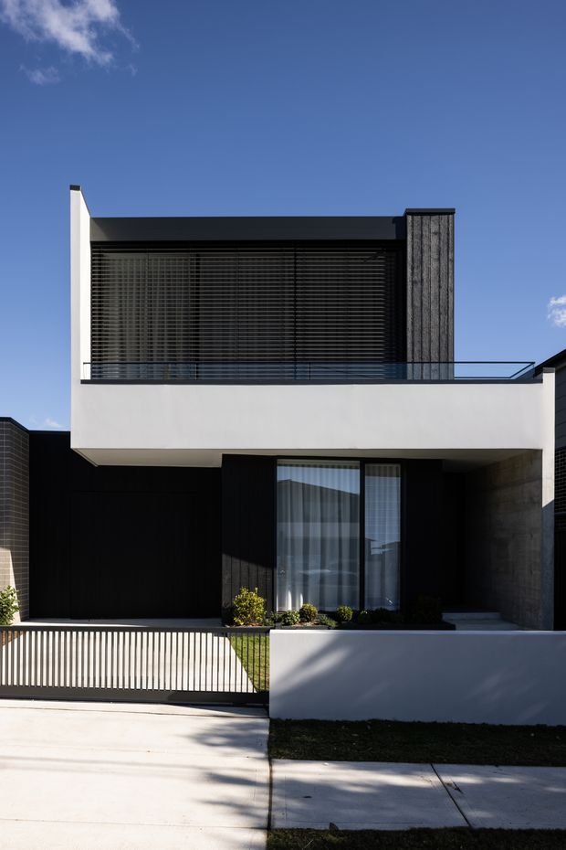 Street-facing view of a contemporary two-storey residence with a crisp geometric composition. The facade combines white rendered planes, dark cladding, extensive upper-level glazing screened by horizontal blinds, a textured vertical feature wall, and a slim glass balustrade. A low front wall, vertical-slat gate, driveway, and small landscaped strip complete the foreground.