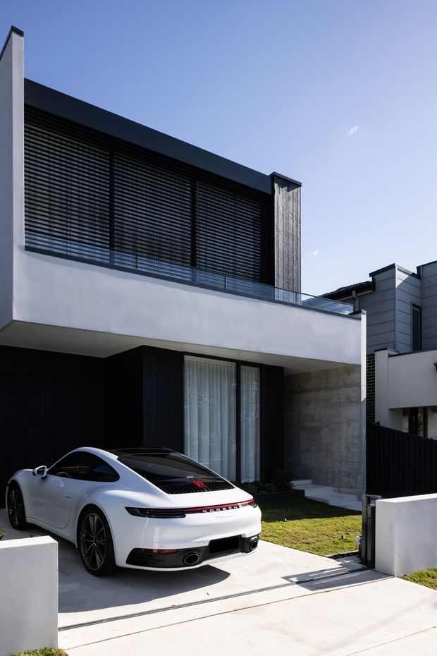 Exterior photograph of a contemporary multi-level residence with a crisp white rendered facade, dark horizontal louvres screening the upper-level glazing, a projecting flat roof, concrete feature wall, glass balustrade, and a recessed ground-floor entry. A driveway and garage/carport area occupy the foreground.