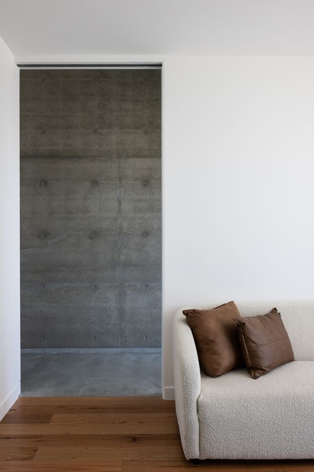 A minimalist interior corner featuring a white plastered wall, a recessed exposed concrete alcove or doorway, and warm timber flooring. A cream boucle sofa with brown leather cushions occupies the right side of the frame. The space is softly lit and sparsely furnished, with a strong contrast between the raw concrete surface and the refined neutral furnishings.