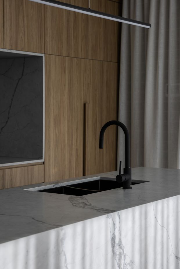 Close-up view of a refined contemporary kitchen island featuring a white veined stone benchtop and matching waterfall edge, an integrated black undermount sink, and a matte black gooseneck tap. The background has warm vertical-grain timber cabinetry with minimal dark pulls, a dark marble-look wall panel, grey curtains, and a slim linear pendant light. The composition is moody, restrained, and materially focused.