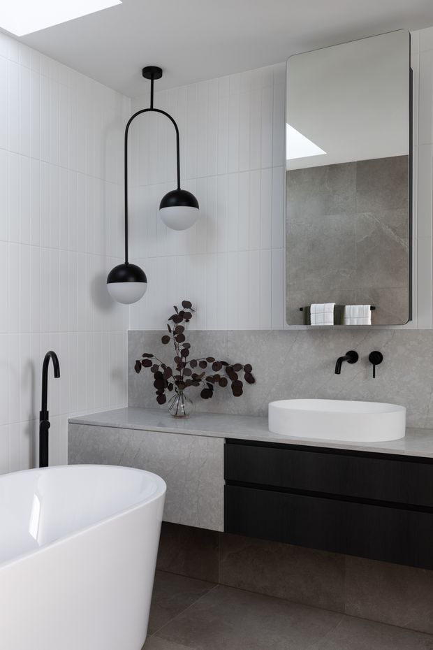 A contemporary bathroom featuring a freestanding white bathtub, floating dark timber vanity with a white vessel basin, wall-mounted black tapware, a large rounded-corner mirror, grey stone-look benchtop and splashback, and white vertically patterned wall tiles. Two black-and-white pendant lights and a skylight provide illumination.