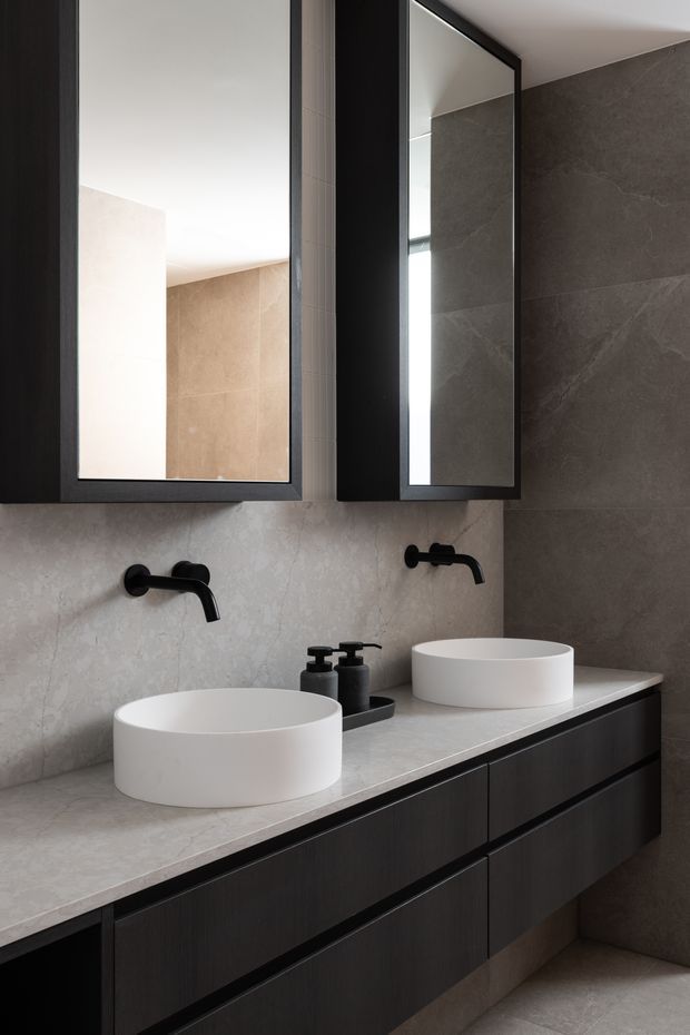 A contemporary double-vanity bathroom with two white round vessel basins on a pale stone benchtop, matte black wall-mounted taps, dark timber-look floating cabinetry, black-framed mirrored cabinets, and large grey-beige stone-look wall tiles. The space has a refined, understated and slightly moody palette.