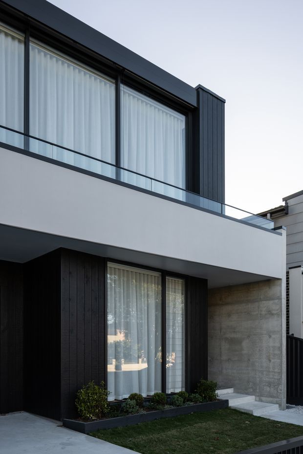 A contemporary two-storey residential facade with a projecting upper-level balcony, large glazed openings, dark vertical timber cladding, smooth white rendered walls, and an exposed concrete side wall. Minimal landscaping and a small lawn sit along the ground-floor frontage. The composition emphasizes clean horizontal and vertical lines, restrained materials, and modern detailing.