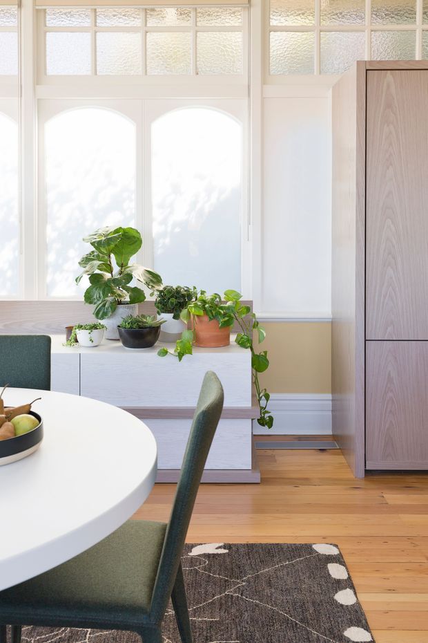 Bright, refined dining area with a round white dining table and upholstered green chairs, positioned in front of tall white decorative glazing and wall panels. A low white built-in sideboard displays potted plants, while a tall light timber storage cabinet stands to the right. Warm timber flooring and a dark patterned rug add contrast to the predominantly light interior.