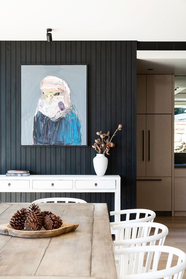A refined dining area with a large pale timber dining table and white spindle-back chairs in the foreground. A white console with drawers sits against a dark vertical timber-panelled feature wall, topped with a ceramic vase and dried branches. Above it hangs a colourful abstract portrait, while warm timber cabinetry and a partially visible kitchen or hallway appear through the opening on the right.