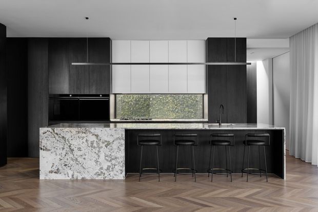 Contemporary kitchen with a large dark timber island and four integrated bar stools, dramatic white-and-grey stone benchtops, dark full-height cabinetry, white overhead cabinetry, concealed appliances, and a slim linear pendant light. The space has a warm herringbone timber floor, a black sink tap, and soft natural light through a rear window and sheer curtains.