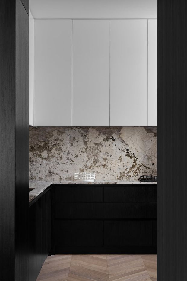 A compact, highly refined modern kitchen viewed through a dark timber-framed opening. The kitchen features matte white upper cabinetry, deep black or dark timber lower cabinetry, a dramatic veined natural-stone backsplash and benchtop, and warm timber herringbone flooring. The concealed, handleless joinery creates a minimalist, tonal composition with strong material contrast.