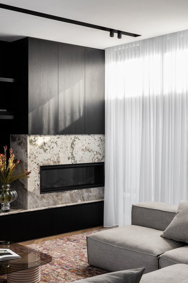 A refined contemporary living room featuring a dark built-in fireplace wall with tall charcoal cabinetry, a pale natural-stone fireplace surround, and black lower joinery. Sheer floor-to-ceiling curtains filter bright daylight beside a large grey sectional sofa, with a patterned rug and sculptural glass coffee table completing the space.