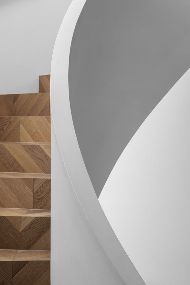 Close-up architectural view of a sculptural staircase with warm herringbone-pattern timber treads and risers, partially concealed behind a sweeping curved white balustrade or wall. The surrounding surfaces are smooth white and soft grey, creating a minimalist, abstract composition.