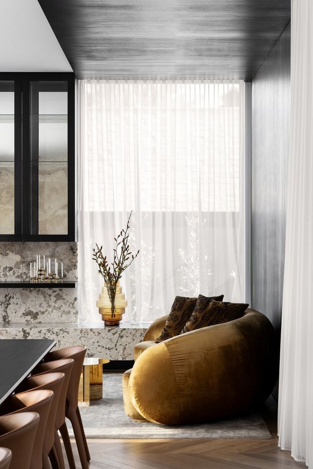 A refined open-plan living and dining area with a sculptural mustard-gold curved sofa, patterned cushions, a dark dining table with upholstered chairs, and a marble-clad fireplace or joinery wall. Sheer white curtains cover a large window, while dark wood or metal paneling wraps the ceiling and side wall. The room has a warm, luxurious, moody contemporary character with herringbone timber flooring and layered natural light.