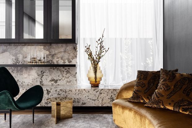 A richly styled contemporary living room with a sculptural dark green lounge chair, a curved gold velvet sofa with patterned cushions, and a small metallic side table. The room features dramatic veined stone across the fireplace or media wall and hearth, dark framed cabinetry, sheer floor-to-ceiling curtains, dark timber wall panelling, and a decorative vase with branches. The palette is moody, warm, and luxurious with strong textural contrast.