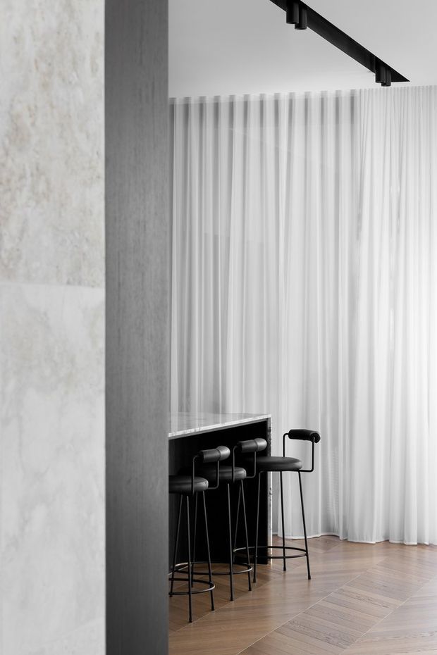 A refined contemporary interior featuring a compact black bar with several high stools, framed by a pale stone or concrete wall and sheer full-height curtains. A black ceiling-mounted track light with cylindrical spotlights contrasts against the light ceiling, while warm timber flooring adds softness to the monochrome scheme.