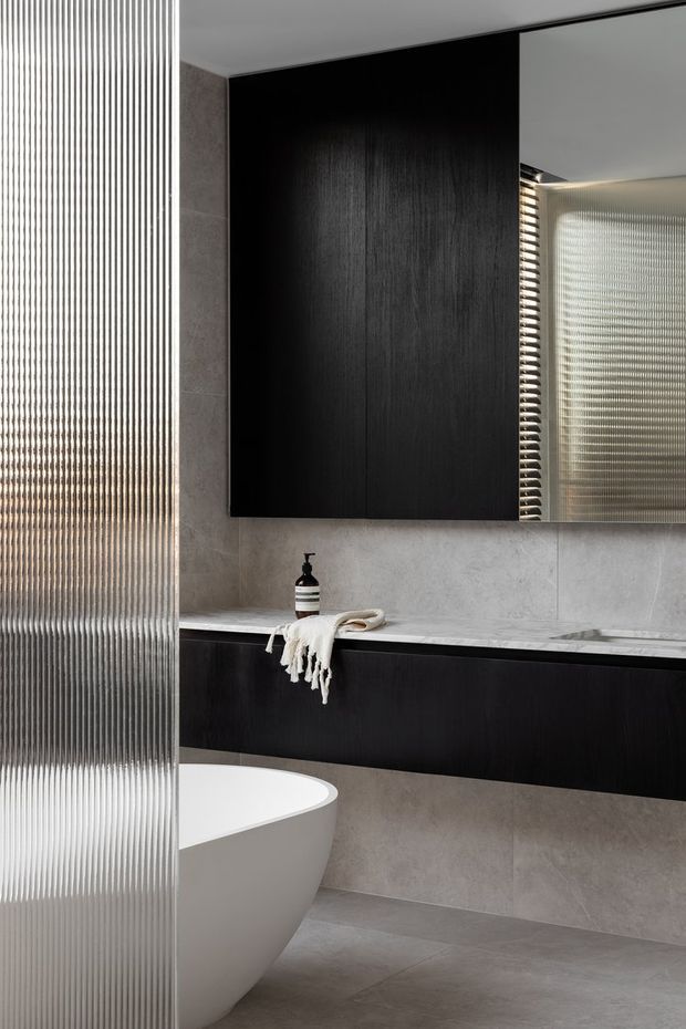 A refined contemporary bathroom featuring a freestanding white bathtub, dark timber-toned wall-hung cabinetry, a large black vanity cabinet, grey stone-look wall and floor surfaces, and a ribbed translucent glass partition. A mirrored cabinet reflects the textured glazing, while soft integrated lighting adds a calm, luxurious atmosphere.