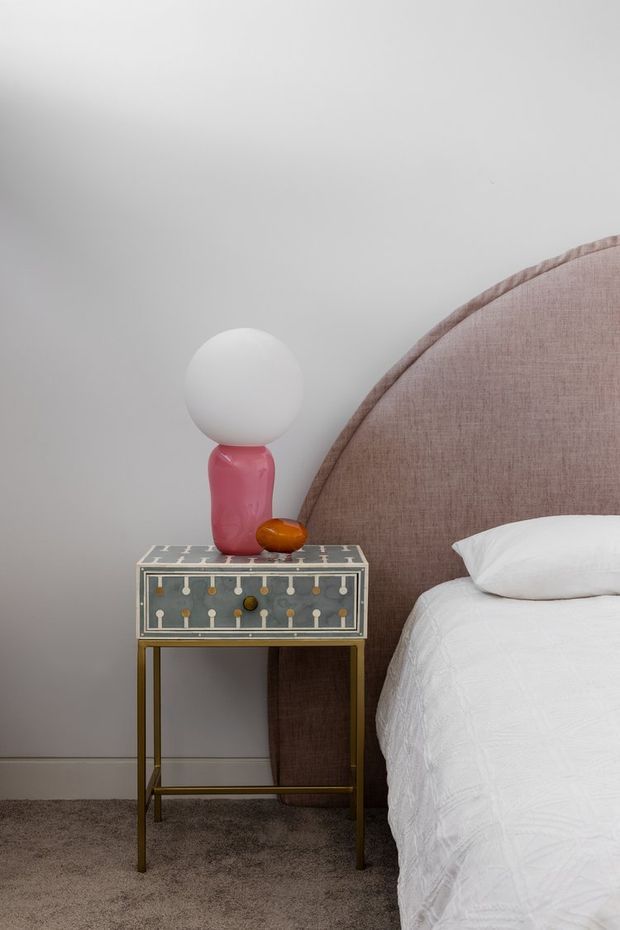 A close-up bedroom vignette featuring a partially visible bed with a large rounded upholstered blush-pink headboard, white bedding and pillow, and a decorative bedside table with patterned drawer fronts and slender brass-toned legs. A sculptural pink and white lamp and small orange accent sit on the table. The room has light grey walls and taupe carpet, creating a refined, softly contemporary atmosphere.