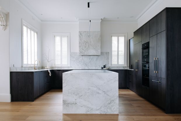A bright, refined kitchen with a large white veined stone island, dark timber or stained-wood cabinetry, matching dark wall-to-wall joinery, white walls, plantation shutters, and warm timber flooring. The layout includes an integrated oven stack, a concealed or integrated rangehood with marble-look surround, and a minimal pendant over the island.