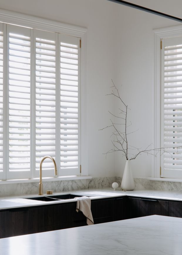 Bright, refined kitchen interior with white plantation shutters, ornate white window trim, marble-look benchtops and splashback, dark timber cabinetry, and a brass gooseneck tap. A sculptural white vase with bare branches provides a minimalist decorative accent.