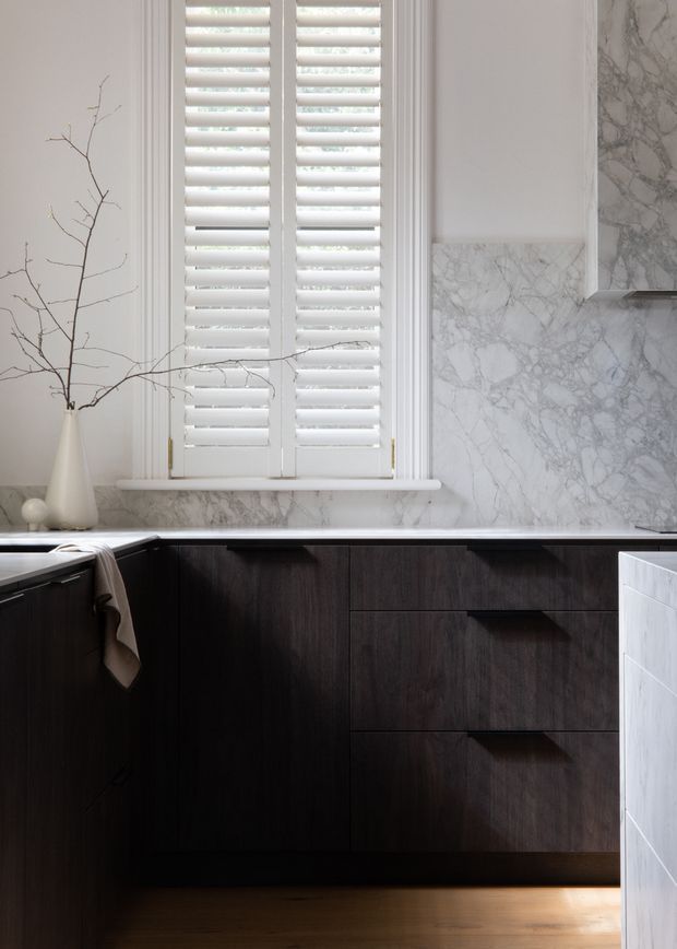 A refined kitchen interior featuring dark timber cabinetry with integrated drawers, a pale veined marble splashback and benchtop, white plantation shutters, and warm timber flooring. The restrained palette and strong contrast create a luxurious, elegant atmosphere.