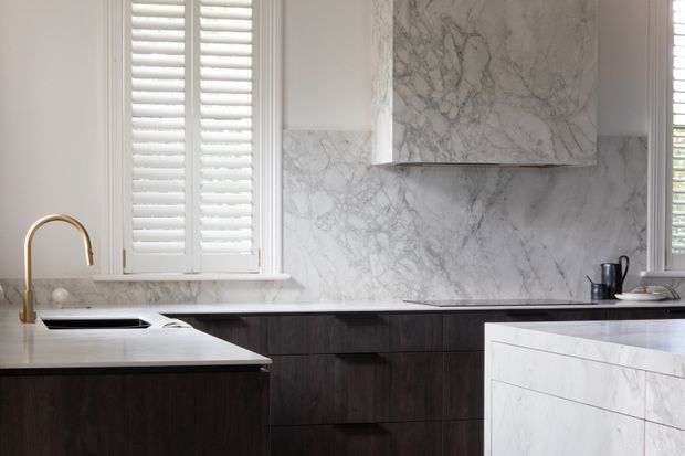 A refined kitchen with dark timber cabinetry, pale veined marble surfaces and splashback, a sculptural marble rangehood surround, white plantation shutters, and a brass gooseneck tap. The composition is clean and high-contrast with a restrained contemporary-classic feel.