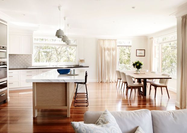 Bright open-plan kitchen and dining area with a white island, integrated cabinetry, subway-tile splashback, timber flooring, pendant lights, and a light upholstered dining setting. Large windows and cream curtains create an airy, refined interior, with part of a living-room sofa visible in the foreground.