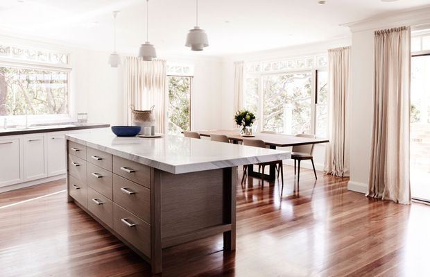 Bright, open-plan kitchen and dining area with a large central island, white perimeter cabinetry, dark wood island joinery, polished light benchtop, timber flooring, pendant lights, and expansive windows dressed with light curtains. A dining table sits beyond the island, surrounded by natural daylight and leafy views.