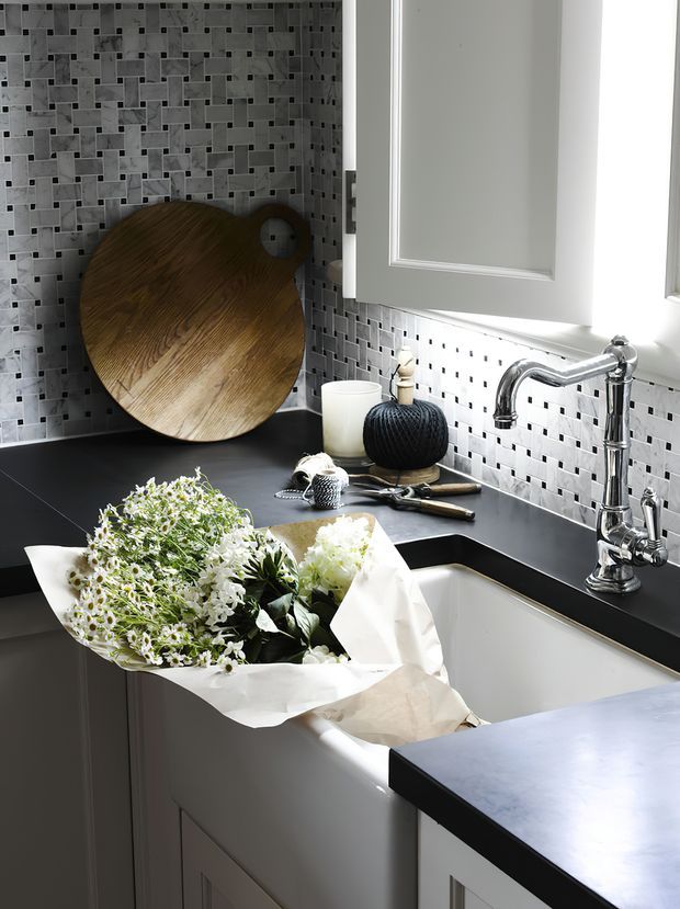 A bright, compact kitchen corner featuring white shaker-style cabinetry, a dark countertop, a white farmhouse-style sink, and a polished chrome bridge faucet beneath a window. The walls are finished with a small grey and white mosaic tile splashback. A round timber cutting board, dark decorative accessories, and a bouquet of flowers sit on the counter.