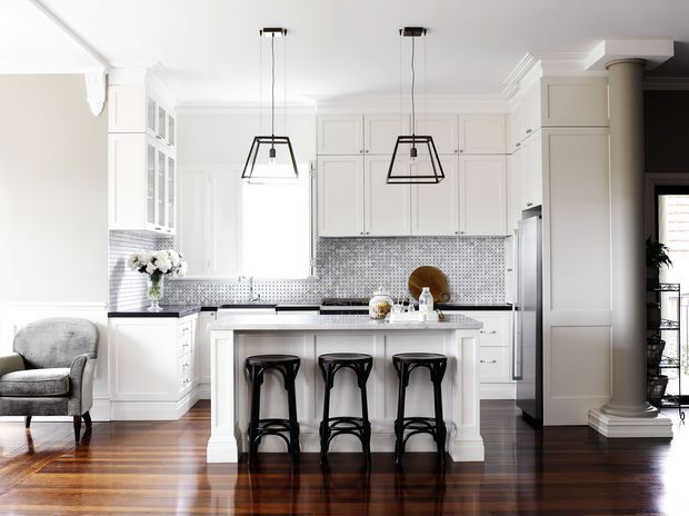 Bright, refined white kitchen with a central island and three black bar stools, surrounded by detailed white cabinetry, a small grey mosaic tile backsplash, dark benchtops, pendant lighting, and polished dark timber flooring. The space opens toward a sitting area and features classical trim, built-in joinery, and a substantial decorative column.