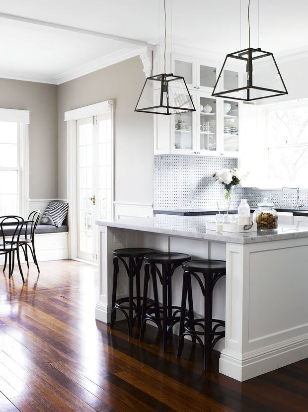 Bright, traditional-style kitchen and dining area with a large white island and grey stone benchtop, seating for several people on black bentwood stools, white cabinetry with glass-front upper cupboards, and a white pegboard splashback. The room opens to a dining nook with a built-in window seat, dark polished timber flooring, extensive white trim and mouldings, and oversized black geometric pendant lights.