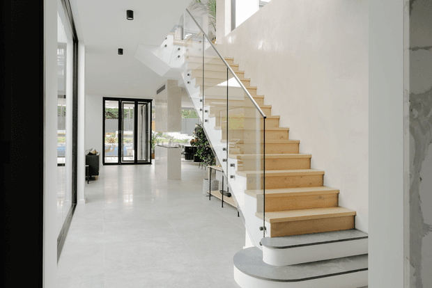 Bright contemporary open-plan interior featuring a sculptural floating timber staircase with a clear glass balustrade and dark grey treads. The staircase rises beside a white double-height wall, with polished light flooring, extensive black-framed glazing, minimalist recessed lighting, and glimpses of living areas and greenery beyond.