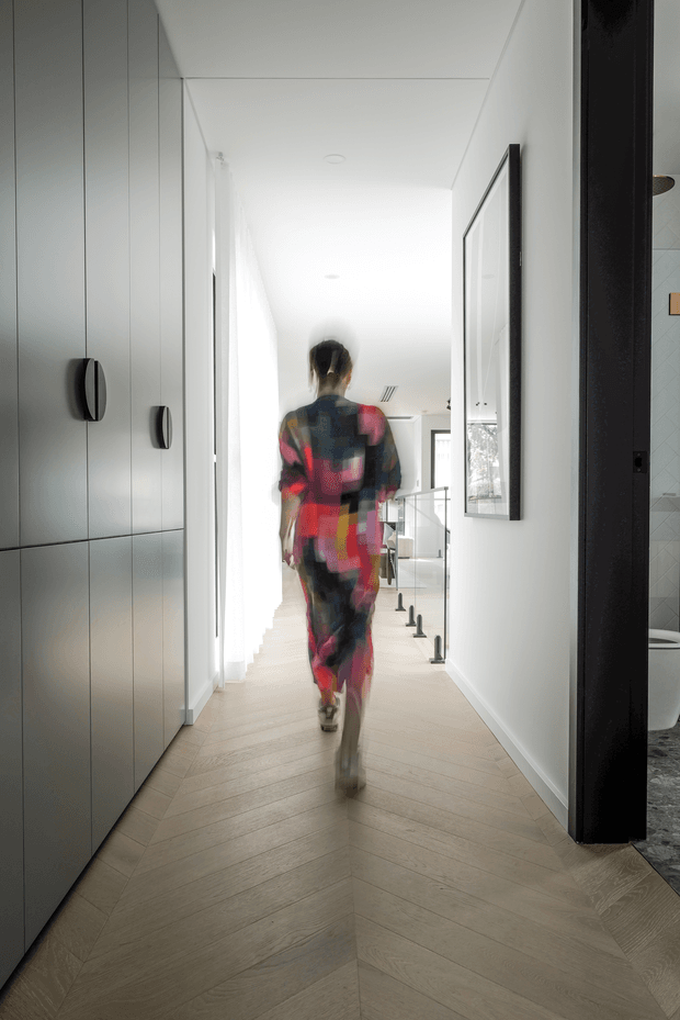 A bright, minimalist residential hallway viewed from inside, with pale herringbone timber flooring, full-height pale built-in cabinetry with dark curved handles on the left, white walls and ceiling, and a large black-framed artwork or mirror on the right. A glass balustrade and stair opening are visible toward the far end, with a bathroom partially visible through a doorway on the right. A blurred person is walking through the corridor.