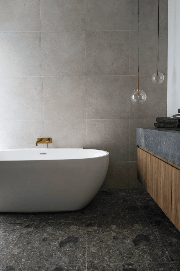 Contemporary bathroom featuring a freestanding white bathtub, large-format grey stone-effect wall tiles, dark textured stone flooring, and a timber vanity with a dark stone benchtop. Two minimalist pendant lights hang beside the tub, creating a refined, moody atmosphere.