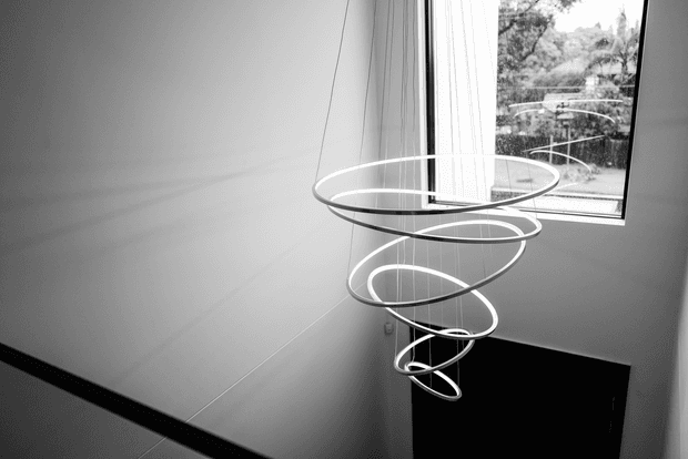A monochrome interior view of a sculptural contemporary pendant light composed of several suspended elliptical rings, hanging in a tall stairwell or vertical circulation space. Smooth pale walls, a dark stair or railing edge, and a large window overlooking trees are visible.
